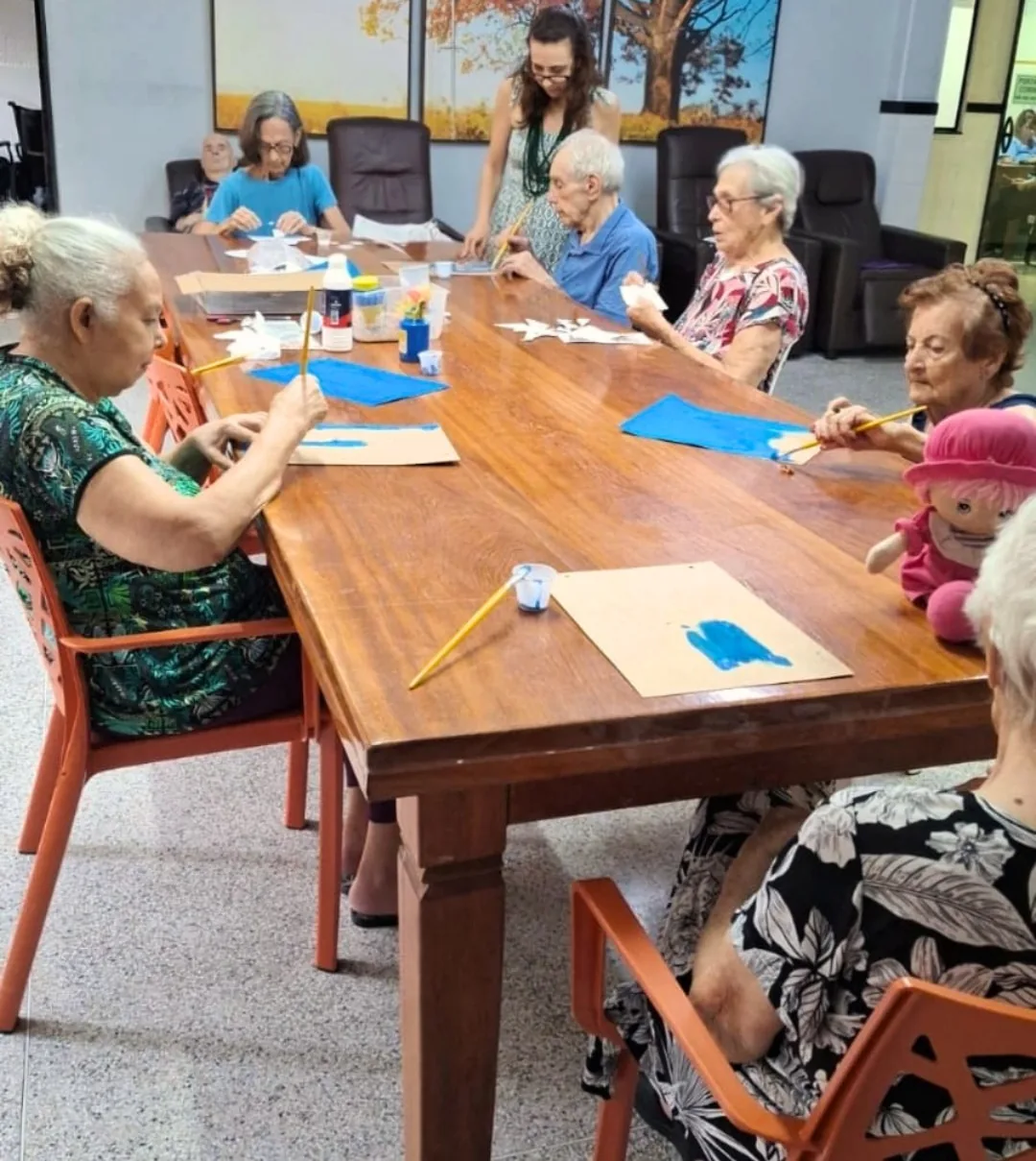 Moradores da LARES pintando em volta da mesa grande da sala, com a terapeuta ocupacional em pé acompanhando