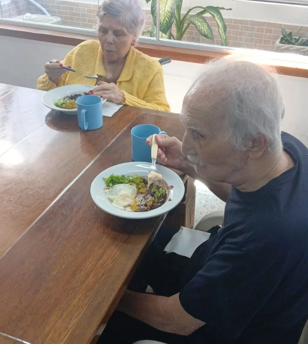Dois moradores da LARES almoçando à mesa de madeira, com prato de arroz, feijão, ovo e salada